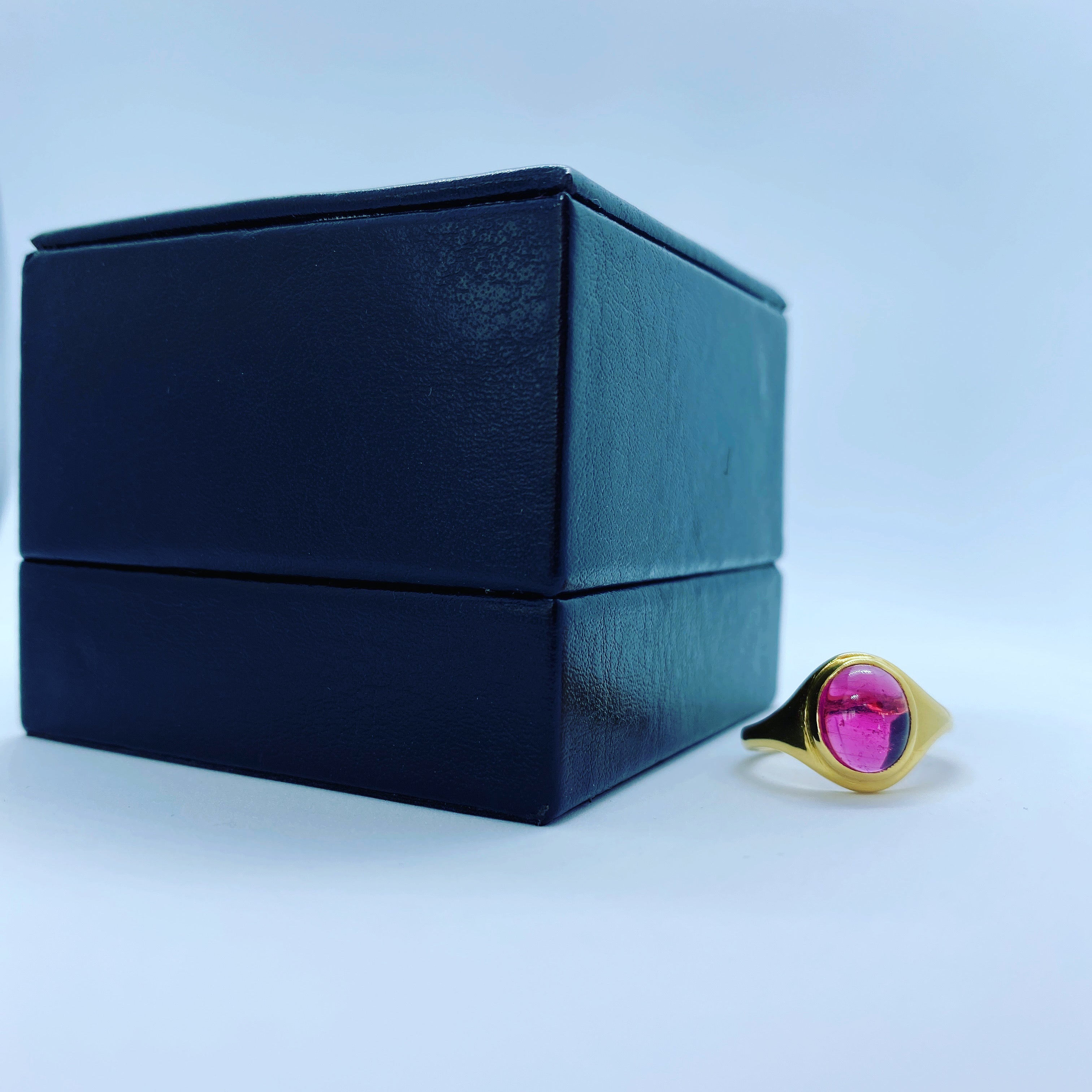A gold signet ring with a vibrant pink oval gemstone, positioned next to a dark navy blue textured jewelry box on a light blue background.