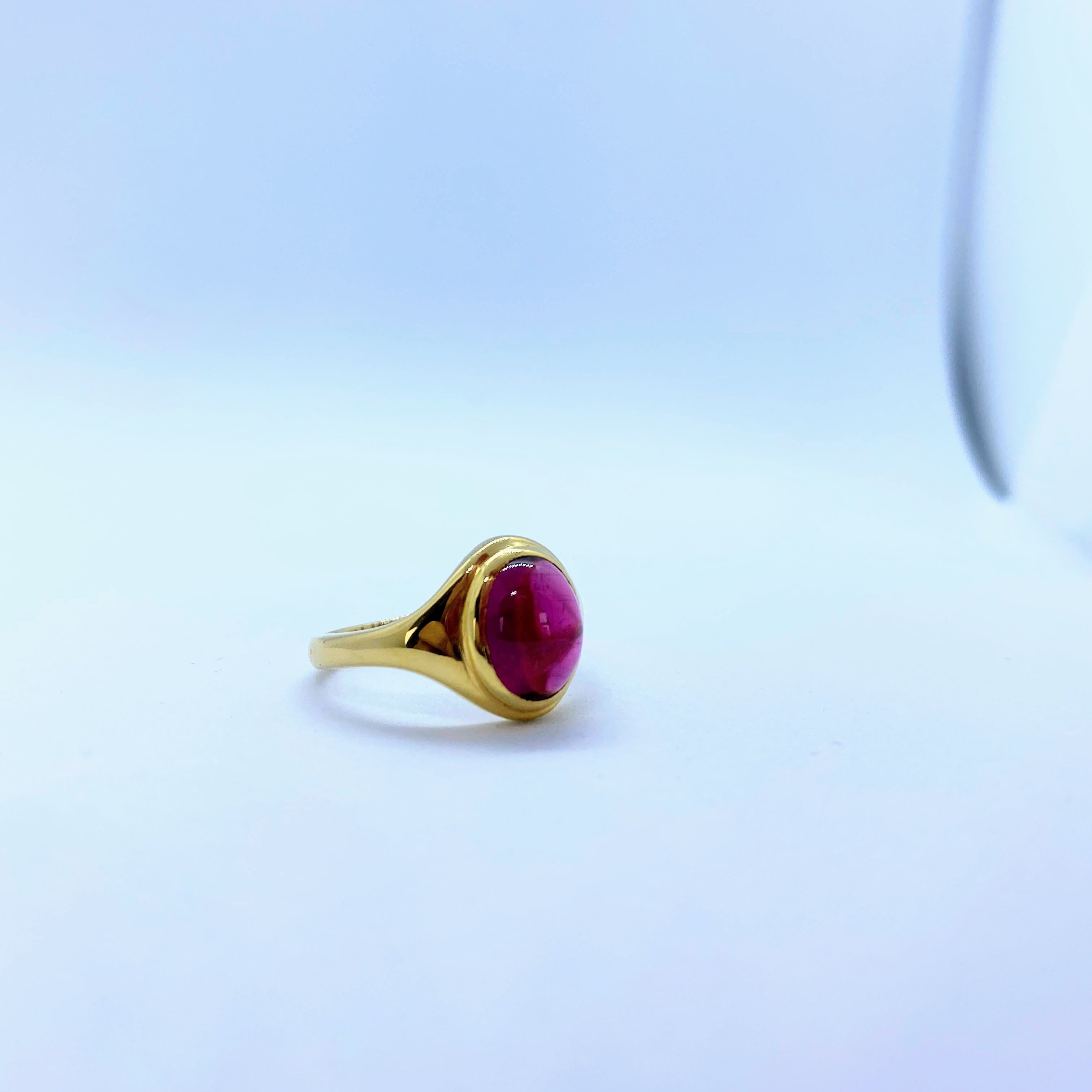 A gold ring with a round, deep red cabochon gemstone, photographed on a light blue and white gradient background.