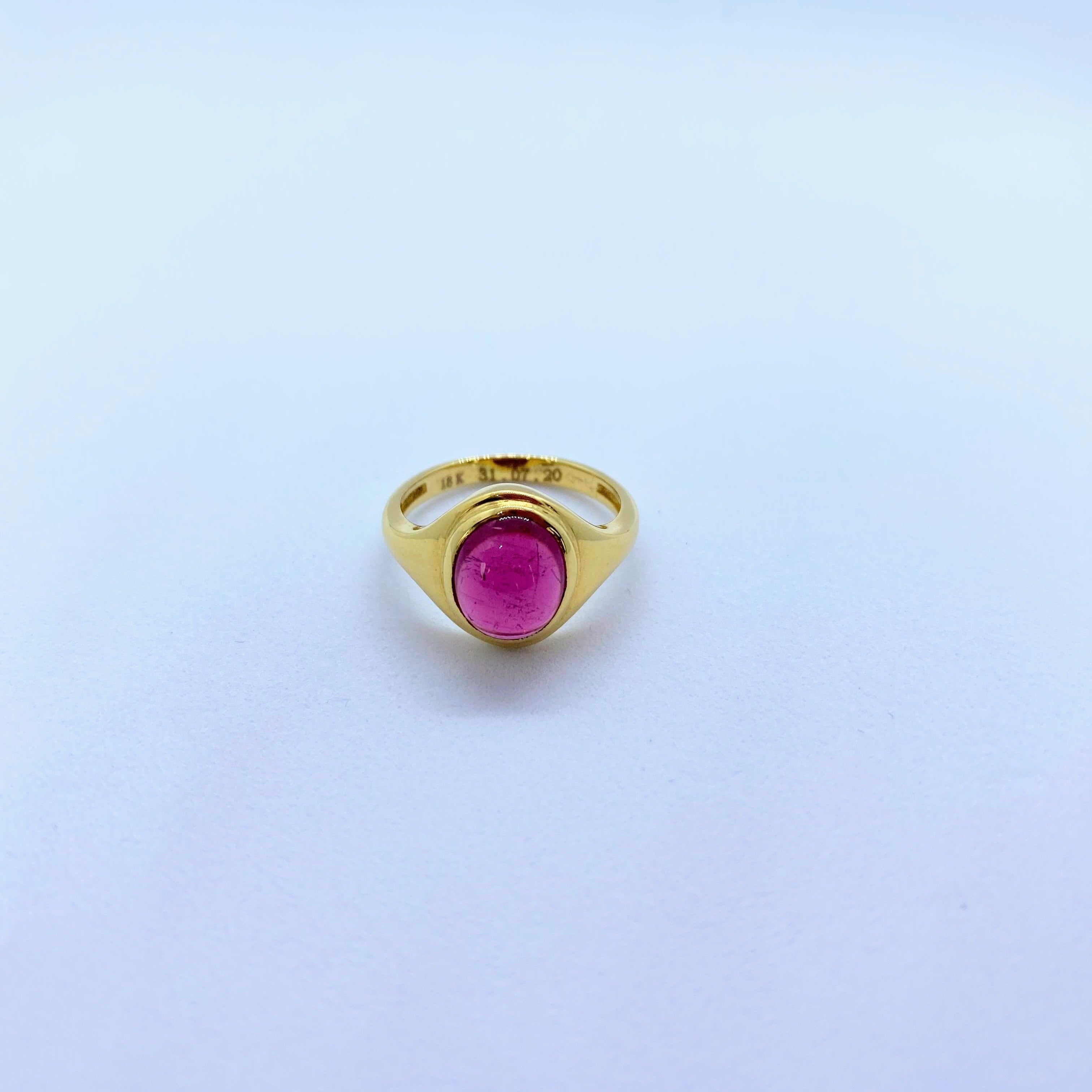 A gold signet ring with an oval pink gemstone, photographed on a light blue gradient background.