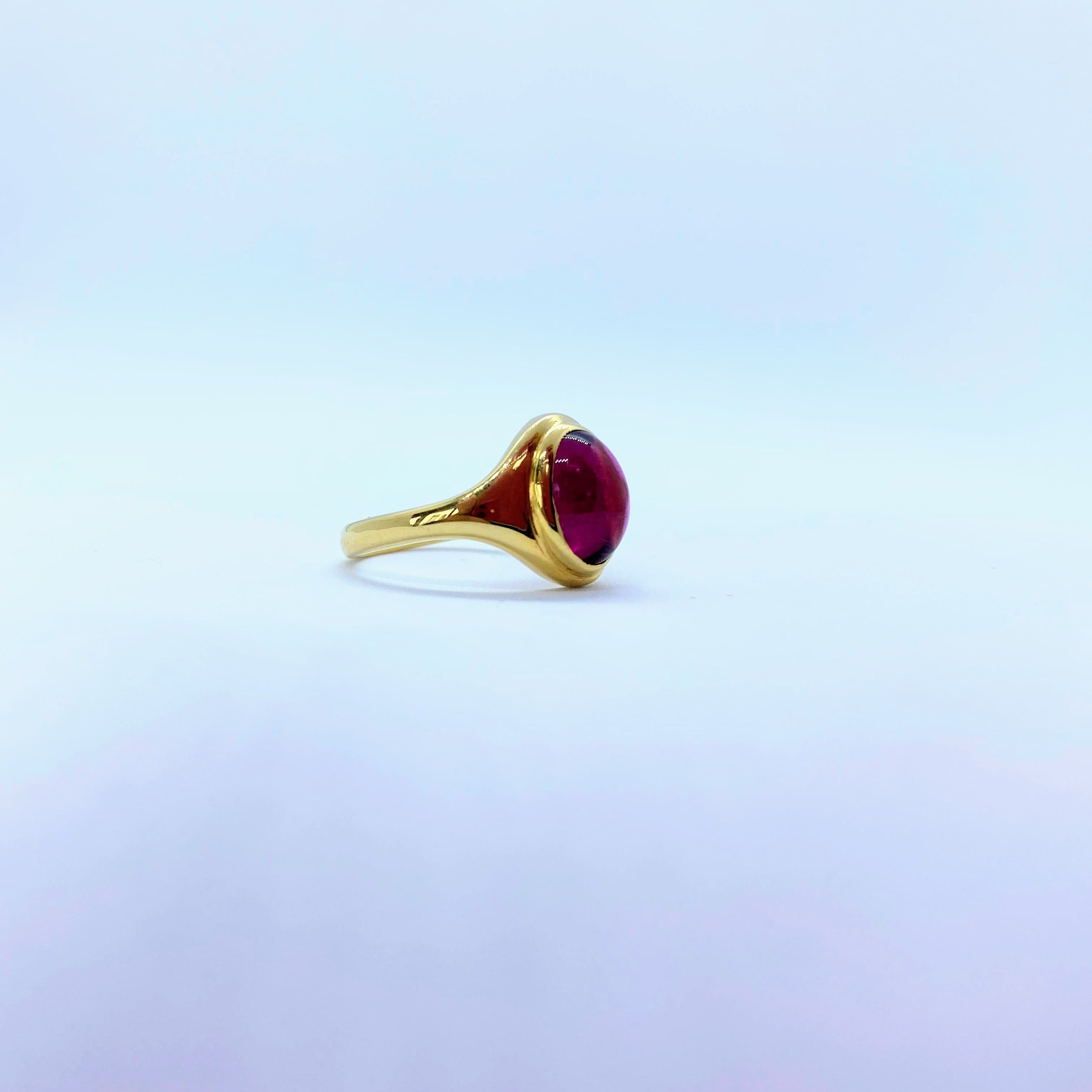 A gold ring with a round, deep red gemstone, photographed on a soft, light blue and lavender gradient background.