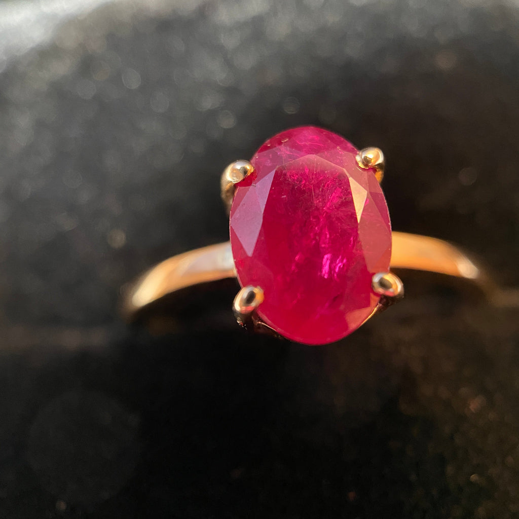 An oval-cut pinkish-red gemstone ring set in a gold band with a four-prong setting, photographed close-up against a dark, textured background.