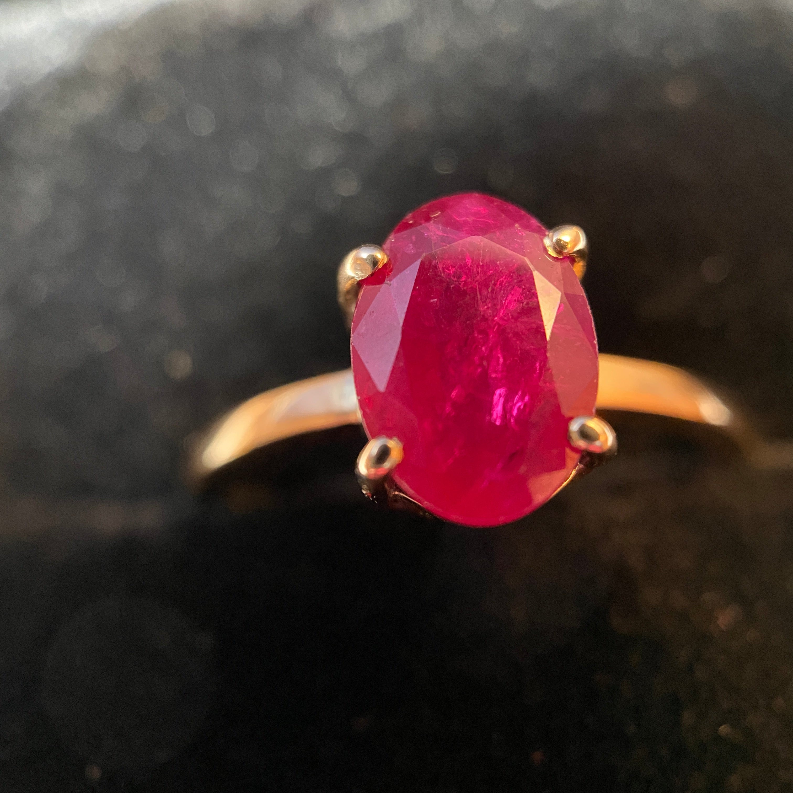 An oval-cut pinkish-red gemstone ring set in a gold band with a four-prong setting, photographed close-up against a dark, textured background.
