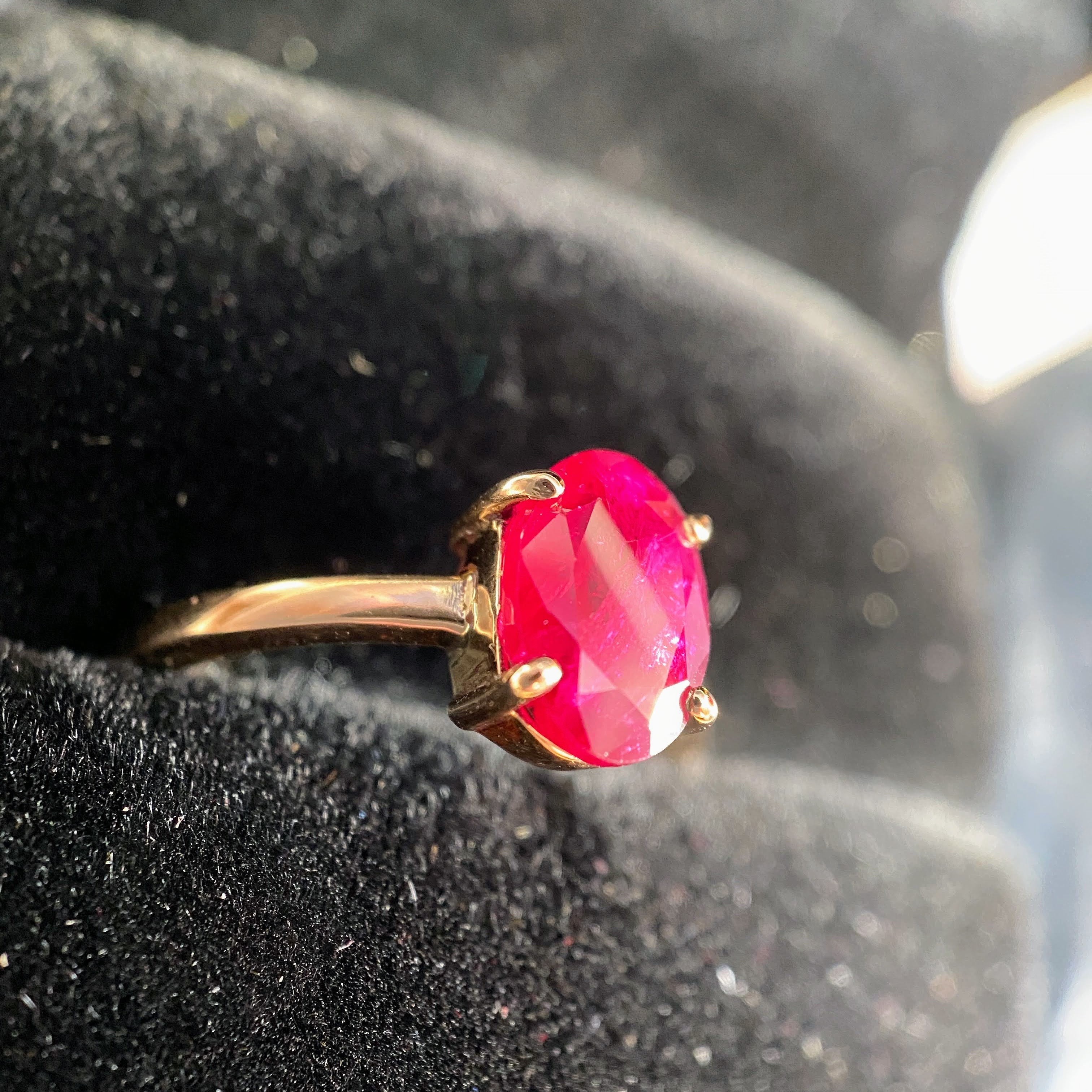 A gold ring with an oval-cut bright pink gemstone, displayed on a textured black velvet ring stand.