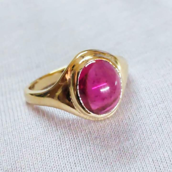 A gold signet-style ring with a round cabochon pink gemstone, resting on a textured white fabric background.